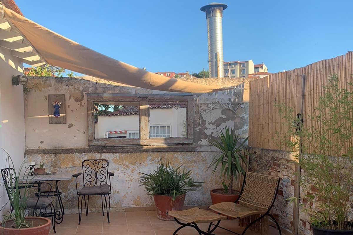Un patio con suelo de baldosas, mobiliario exterior, plantas en macetas y vista a un edificio con chimenea.