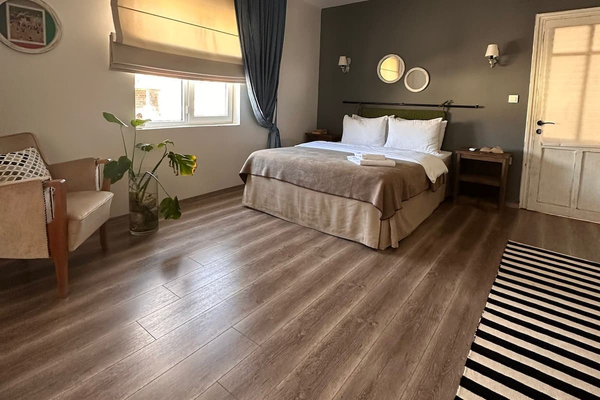Un dormitorio con cama, una silla, una mesita de noche, cortinas en la ventana, una planta en maceta y una alfombra a rayas.