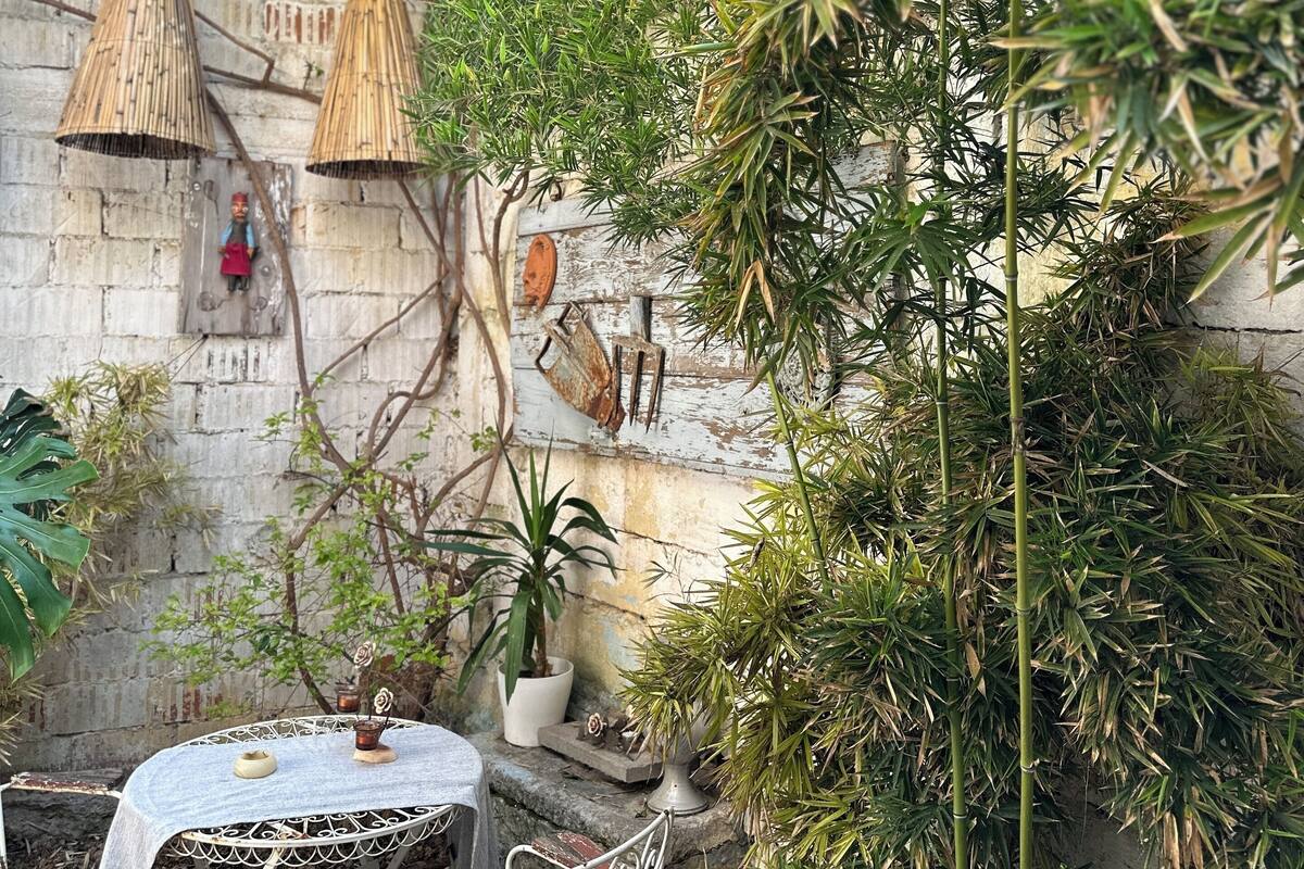 Un espacio de descanso rústico al aire libre con una mesa y sillas de metal, rodeado de exuberante vegetación y un muro de piedra.