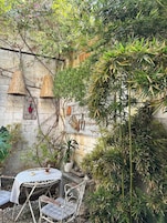 Un espacio de descanso rústico al aire libre con una mesa y sillas de metal, rodeado de exuberante vegetación y un muro de piedra.