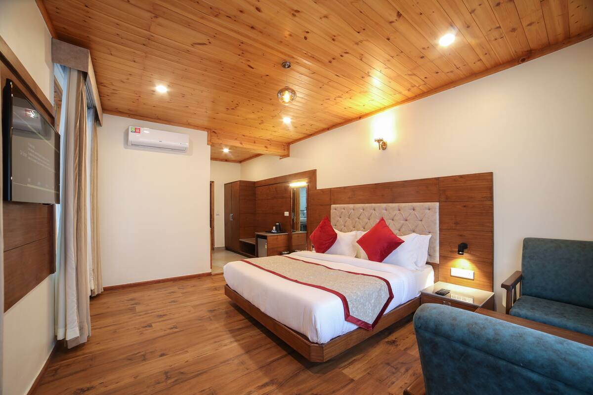 Habitación de hotel con techo de madera, cama con ropa de cama roja y blanca, mesita de noche con lámpara y un sillón azul.