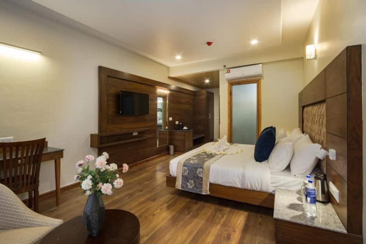 Habitación de hotel con piso de madera, cama con ropa blanca, cabecera de madera, mesita de noche con una botella de agua, un florero con flores y un televisor montado en la pared.