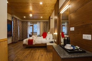 Quarto de hotel com teto de madeira, cama com roupa de cama branca e almofadas vermelhas, mesa de cabeceira com xícara e pires, e vista para o exterior pela janela.