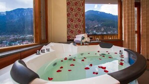 Une salle de bain avec une grande baignoire remplie d’eau et de pétales de rose, une fenêtre donnant sur les montagnes et un rideau à motifs.