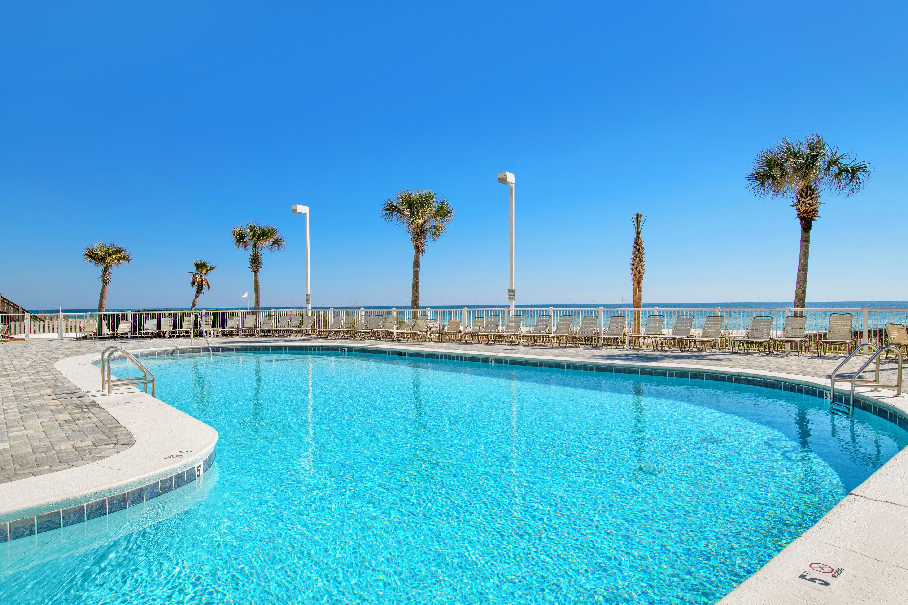 A clear blue pool with a tiled edge, surrounded by lounge chairs and palm trees, with a clear sky above.
