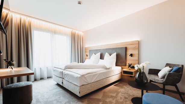 A modern hotel room with a large bed, a desk, a chair, and a small table. - Hotel VIVO (Schierling)