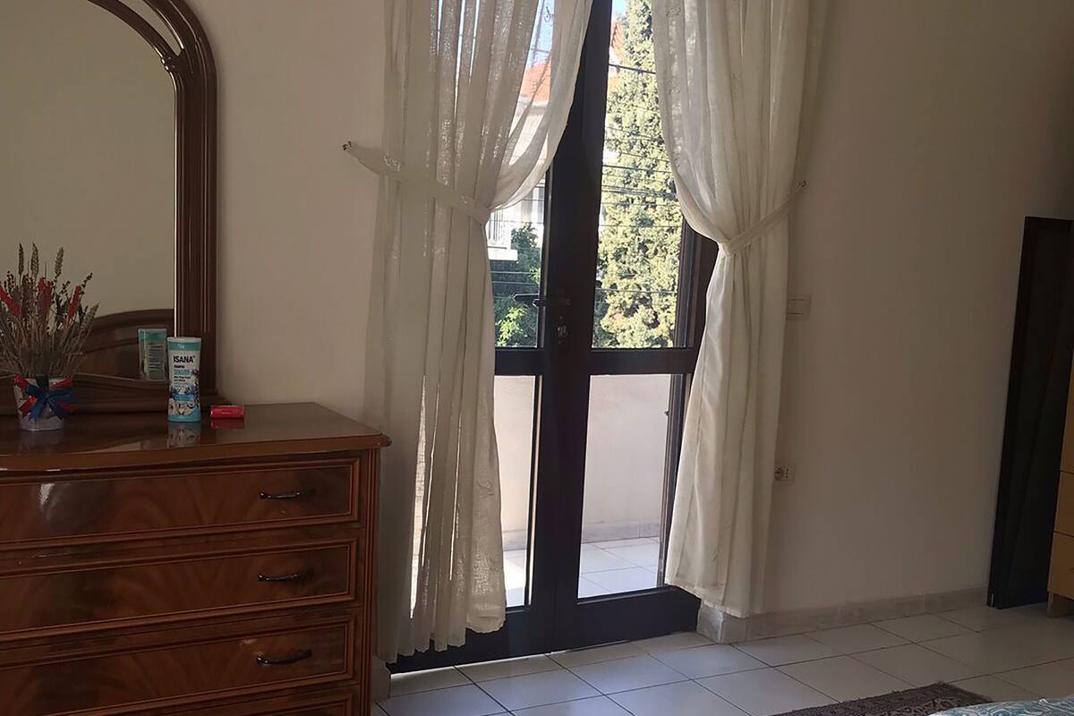 Een slaapkamer met een houten dressoir, een grote spiegel, een bed met een decoratieve spreideken, en uitzicht door een raam met dunne gordijnen.