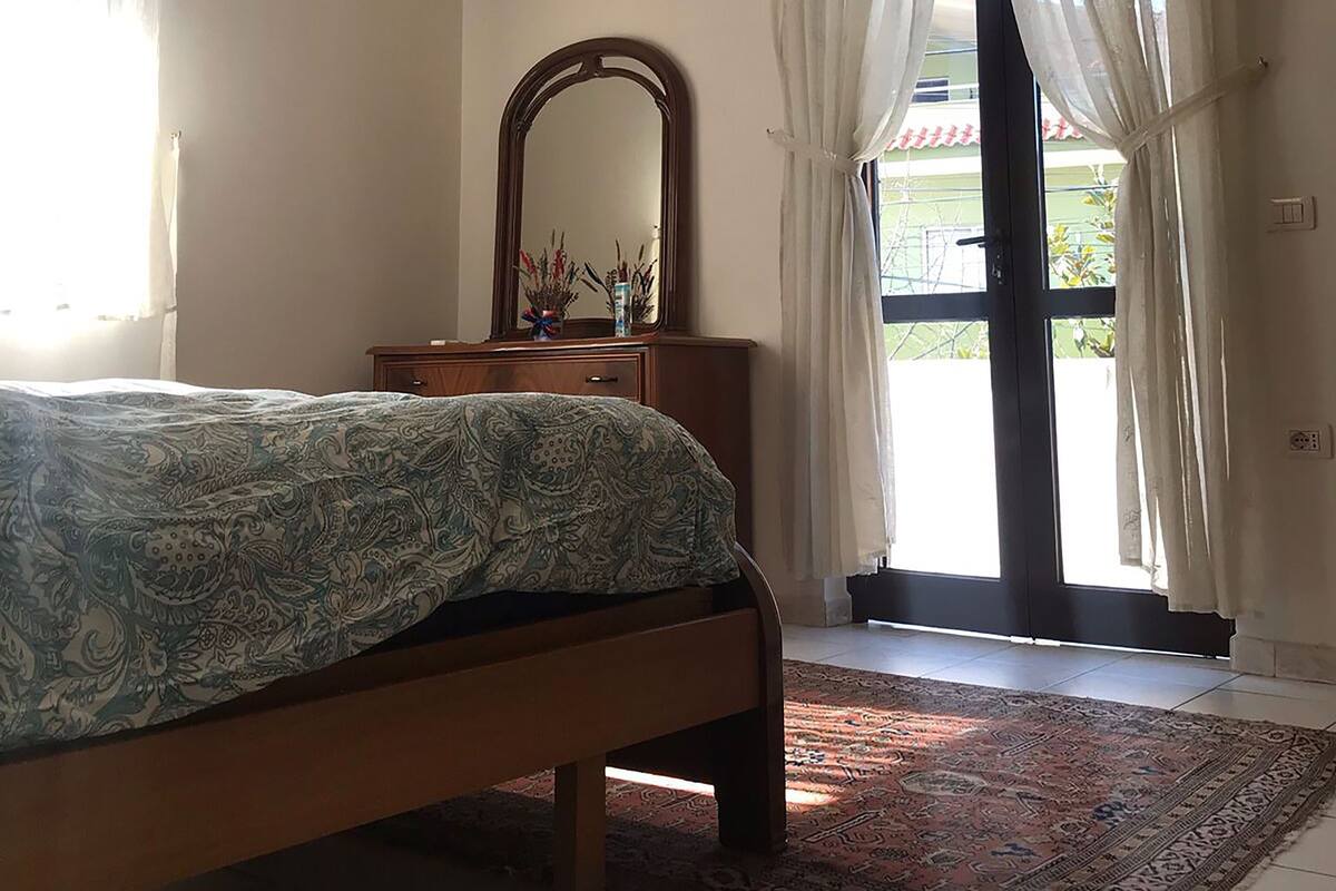 Een slaapkamer met een bed, een dressoir, een spiegel en een raam met gordijnen.