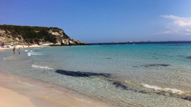 Una playa con agua turquesa cristalina, orilla arenosa y acantilados rocosos al fondo.