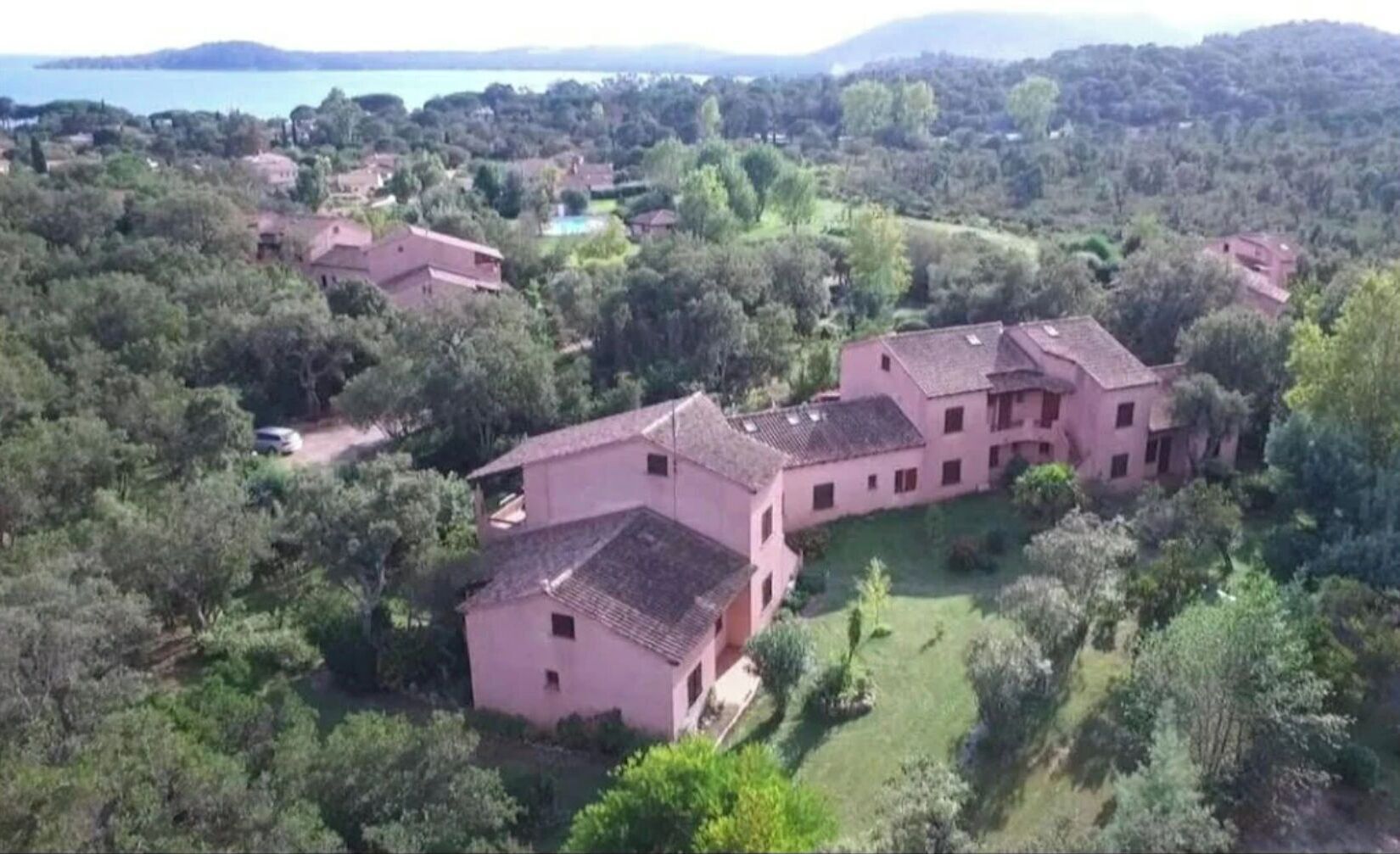 A large, pink villa with multiple buildings, surrounded by lush greenery and a lake in the background.