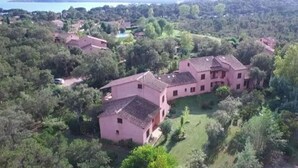 Una amplia villa rosa con varias construcciones, rodeada de exuberante vegetación y un lago al fondo.