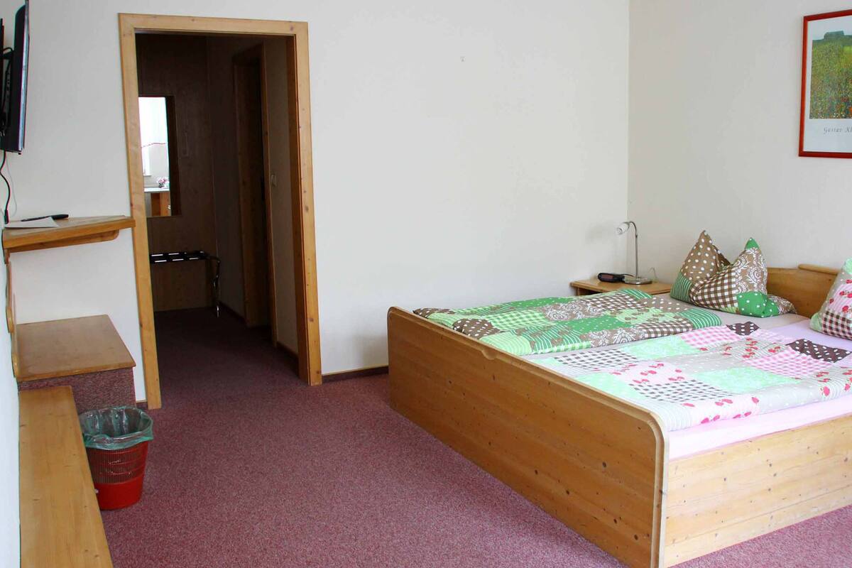 A bedroom with a wooden bed, a desk, and a chair. There is a red trash bin and a wooden shelf.