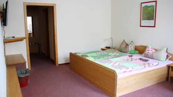 Een slaapkamer met een houten bed, een bureau en een stoel. Er staat een rode prullenbak en een houten plank.