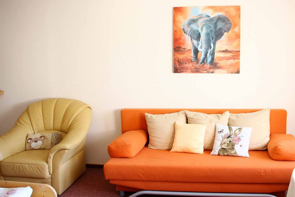 A room with a yellow armchair, an orange sofa with patterned cushions, and a painting of elephants on the wall.