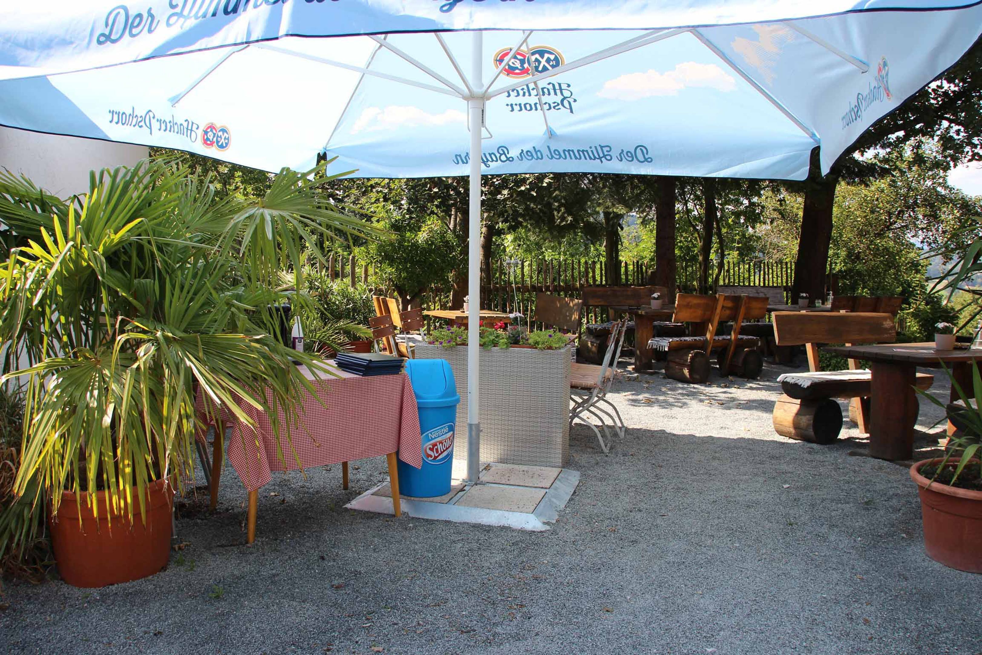 An outdoor seating area with tables and chairs under a large umbrella, surrounded by potted plants and a wicker fence.