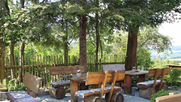An outdoor seating area with rustic wooden furniture, including benches and a table, surrounded by trees and a wooden fence. - Hotel Gasthof Zum weissen Lamm (Hohenberg a.d.Eger)