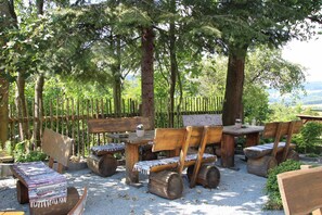 Un espace extérieur aménagé avec des meubles en bois rustique, notamment des bancs et une table, entouré d’arbres et délimité par une clôture en bois.