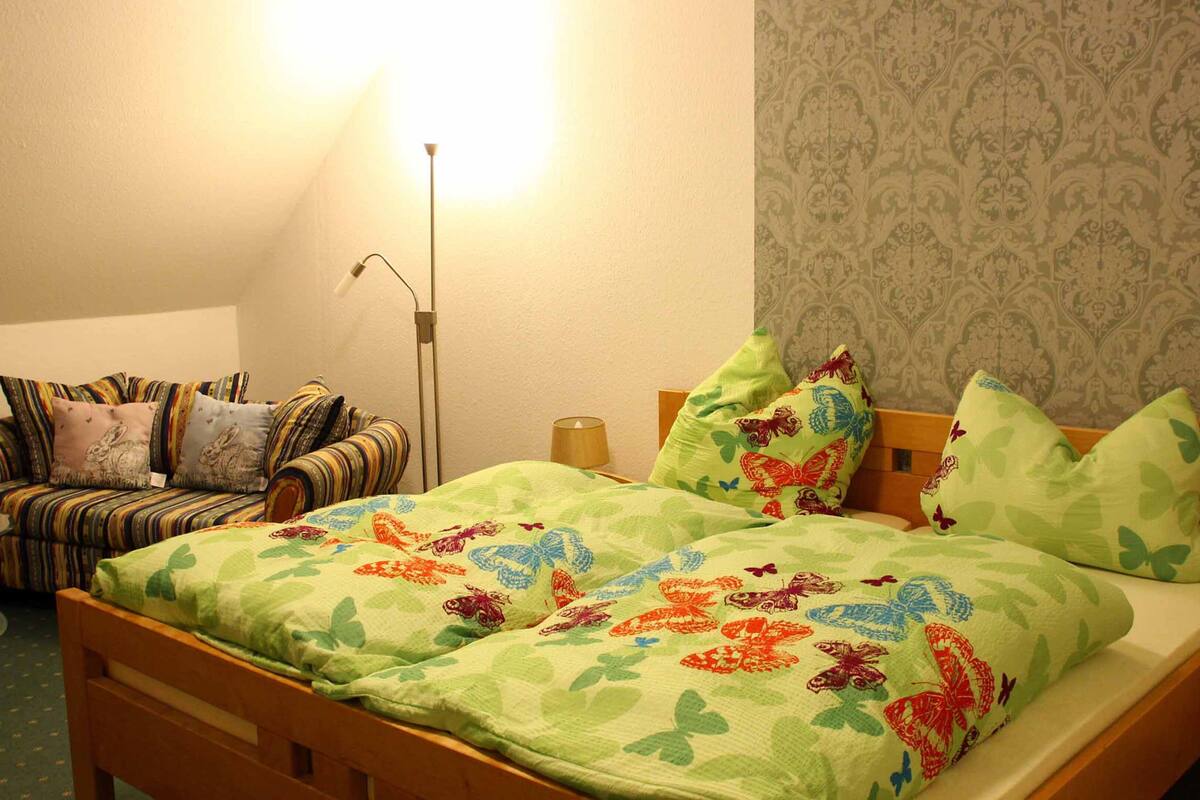 A bedroom with a bed, a sofa, a lamp, and a patterned wallpaper.