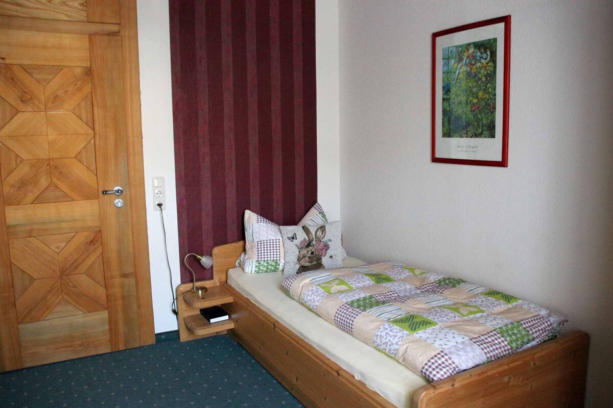 A small bedroom with a wooden bed, a red curtain, a framed picture on the wall, and a wooden door.