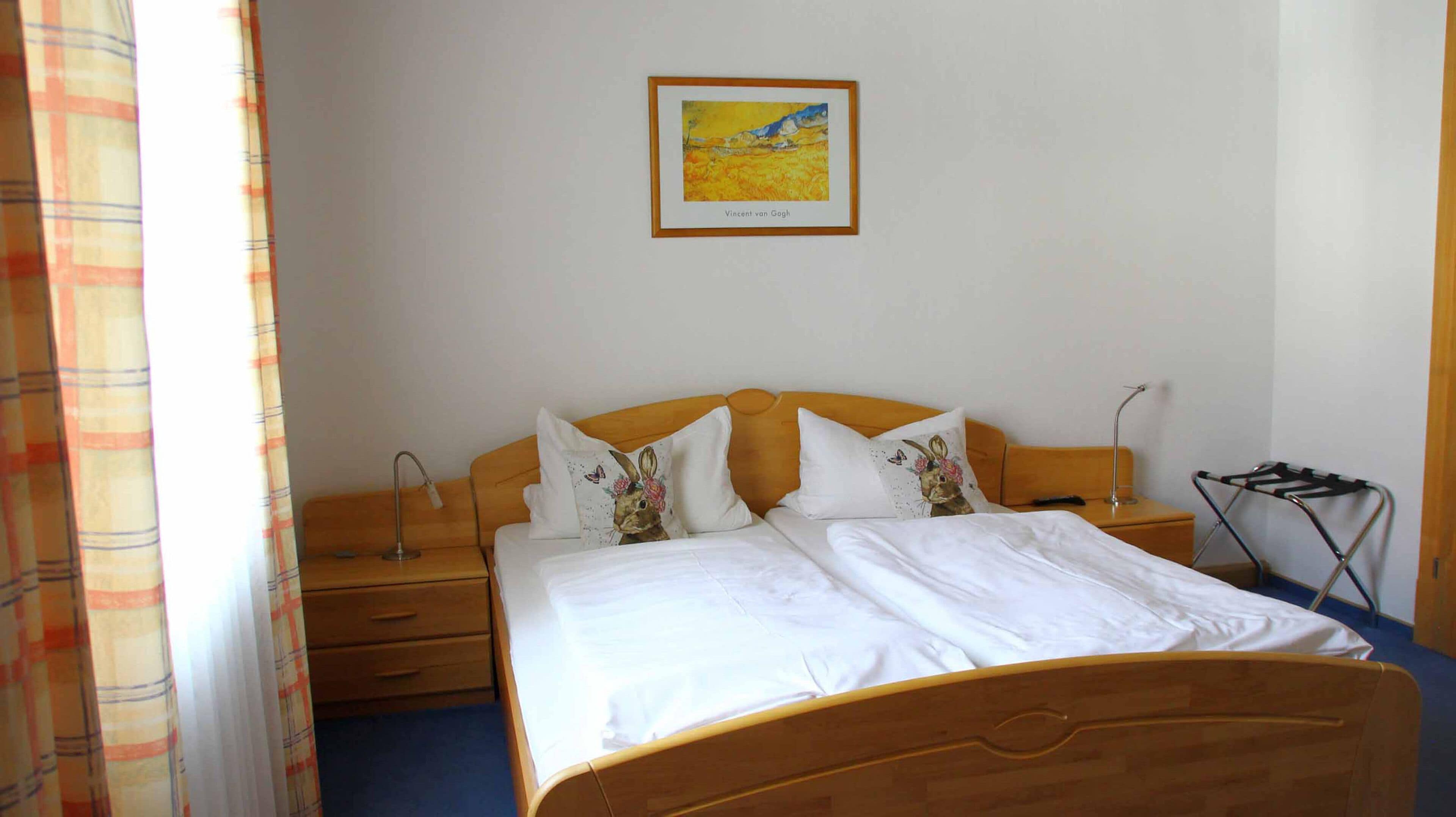 A neatly made bed with white linens, a wooden headboard, and decorative pillows. There is a framed artwork on the wall above the bed, a bedside table with a lamp, and a folding chair.