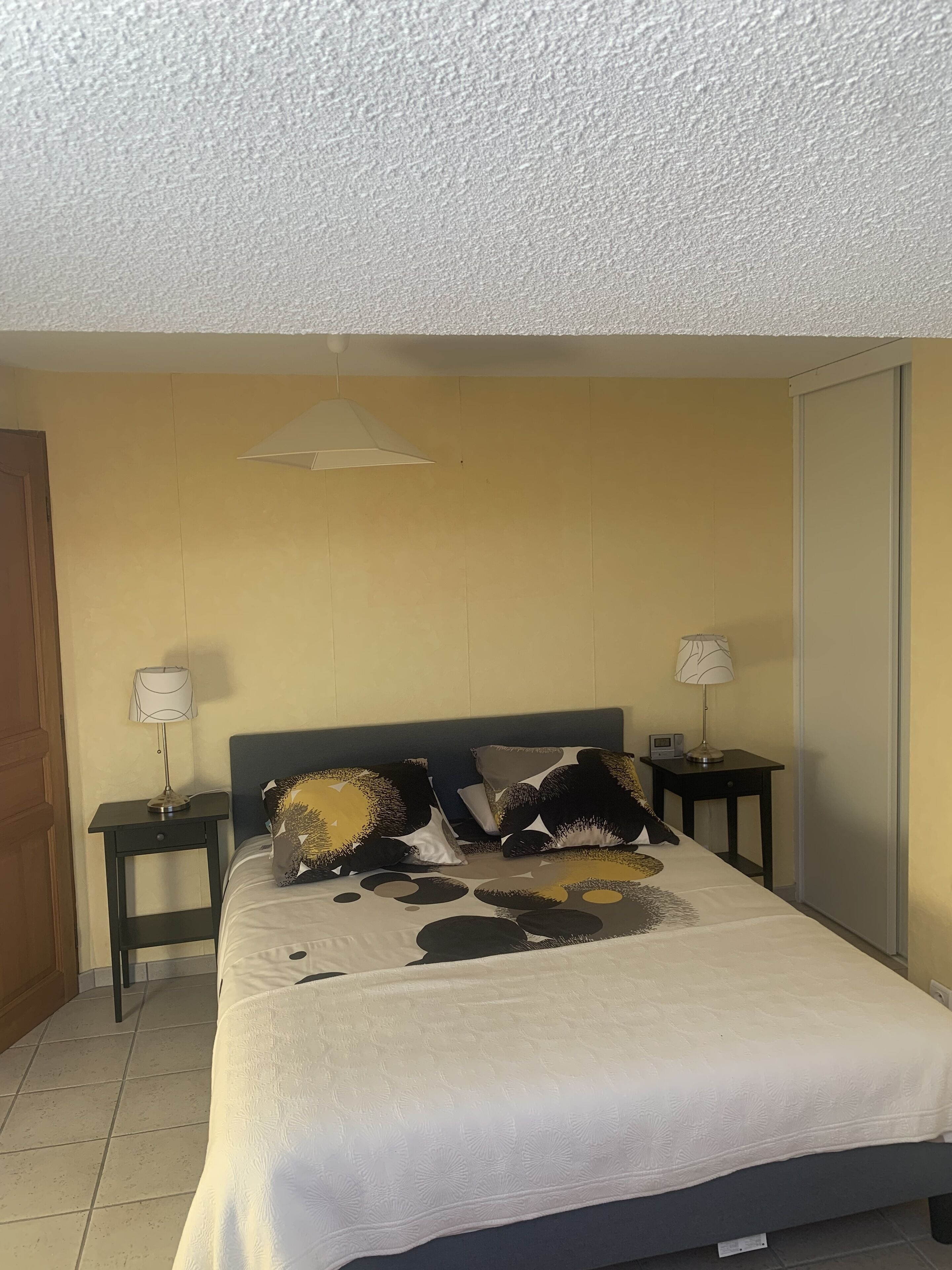 A bedroom with a bed, two bedside tables, a nightstand, a lamp, a wall-mounted light fixture, and a door.