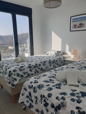A bedroom with two beds, a window with a view of the landscape, and a bedside table with a lamp.