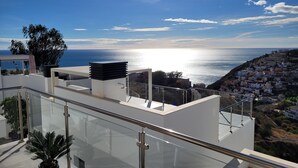 A modern rooftop terrace with a glass railing, a potted plant, and a view of the sea and a coastal town. - Apartment with sea and mountain view. HEATED POOL DURING THE WINTER MONTHS  (Nerja)