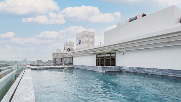 Ein Infinity-Pool auf dem Dach mit Glasgeländer, ein modernes Gebäude mit geometrischen Designelementen und einem wolkenlosen Himmel.