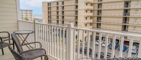 A balcony with wicker chairs and a glass table, overlooking a parking area and a multi-story building with balconies.