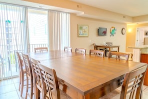 Un comedor con una gran mesa de madera y sillas, una zona de cocina con un microondas y un reloj, y un vidrio corredizo que da a un balcón exterior.
