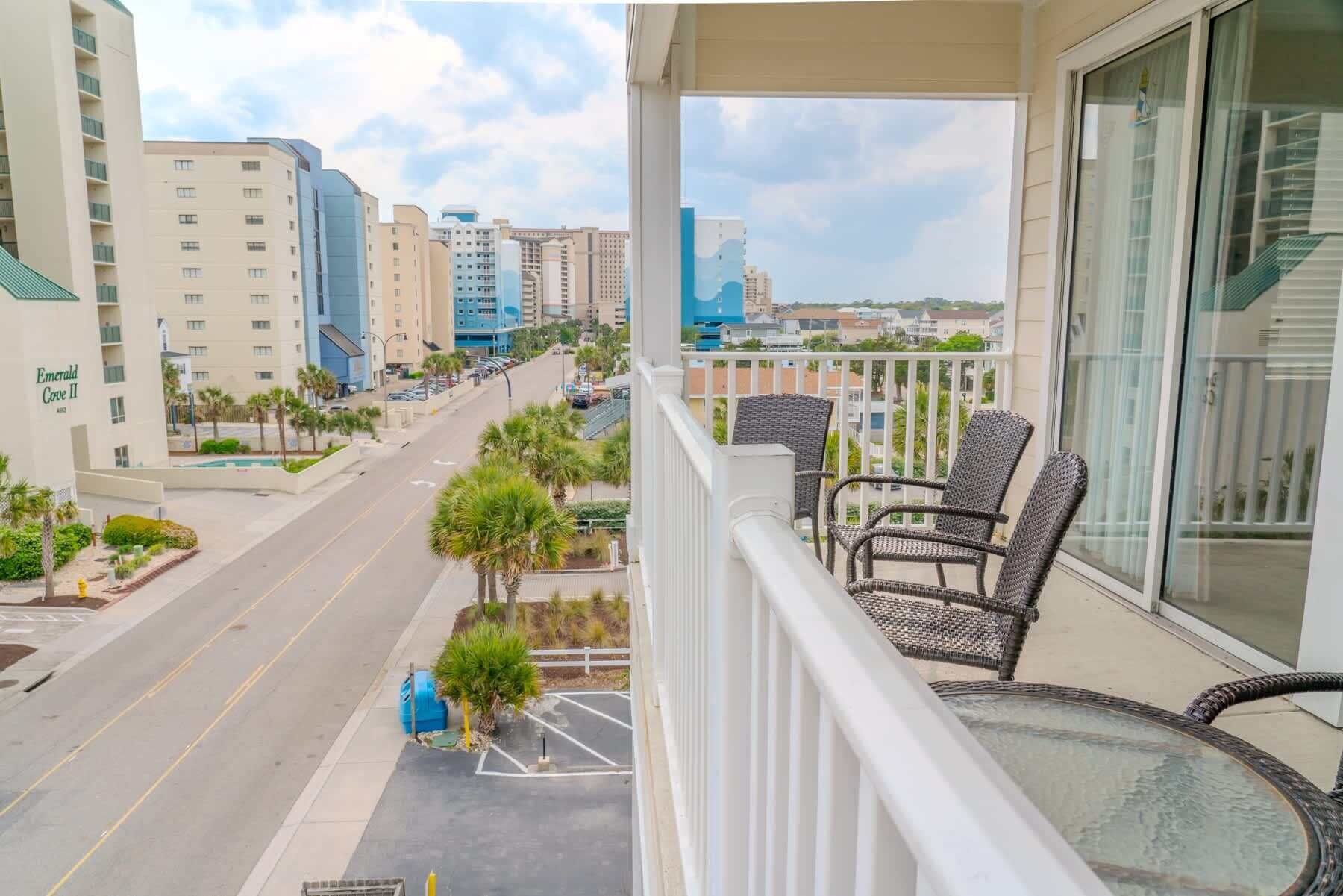 A balcony with wicker chairs, a glass table, and a view of a city street with buildings and palm trees.