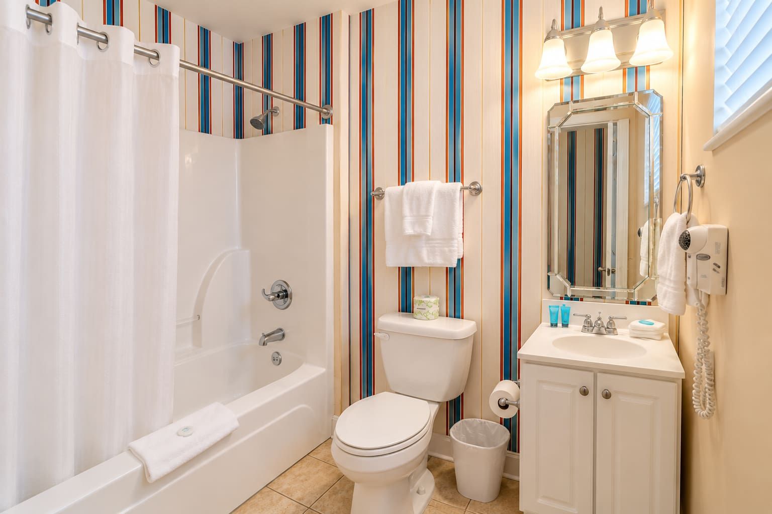 Ein Badezimmer mit Badewanne, Toilette und Waschbecken, ausgestattet mit Streifenmuster-Tapete und einem beleuchteten Spiegel.