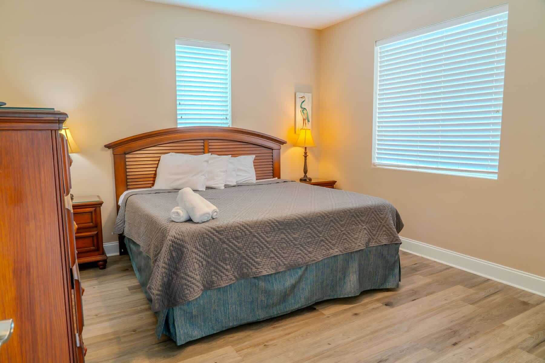 A bedroom with a wooden bed, two bedside tables, a lamp, and two windows with blinds.