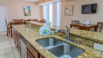A kitchen with granite countertops, a double sink, wooden cabinets, a dining area with chairs, and a TV mounted on the wall.