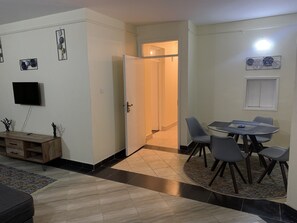 Uma sala de jantar com uma mesa redonda e cadeiras, uma TV fixada na parede e um armário de madeira com gavetas.