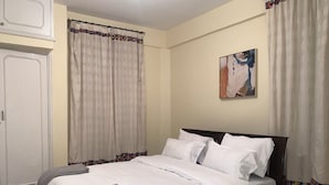 A neatly made bed with white linens and a gray throw blanket, a painting on the wall, and a window with a patterned curtain. - Immaculate 2-bed Apartment, Langata, Nairobi (Nairobi)