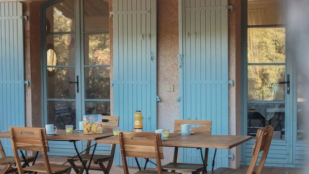 A wooden table with chairs and a box of cookies outside a building with blue shutters and a tiled roof.