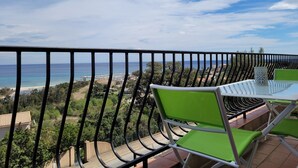 Un balcon avec vue sur la mer et la plage, des chaises vertes et une table.