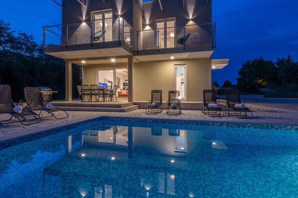 Una casa de dos pisos con piscina, mobiliario exterior y un balcón.