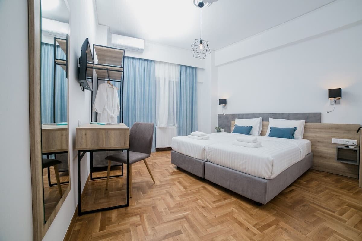 A modern hotel room with a wooden herringbone floor, a desk with a chair, a bed with two pillows, and a wall-mounted TV.