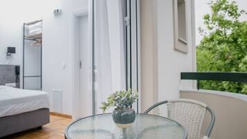 A modern hotel room with a glass table, a potted plant, a bed, and a balcony with a view of greenery.