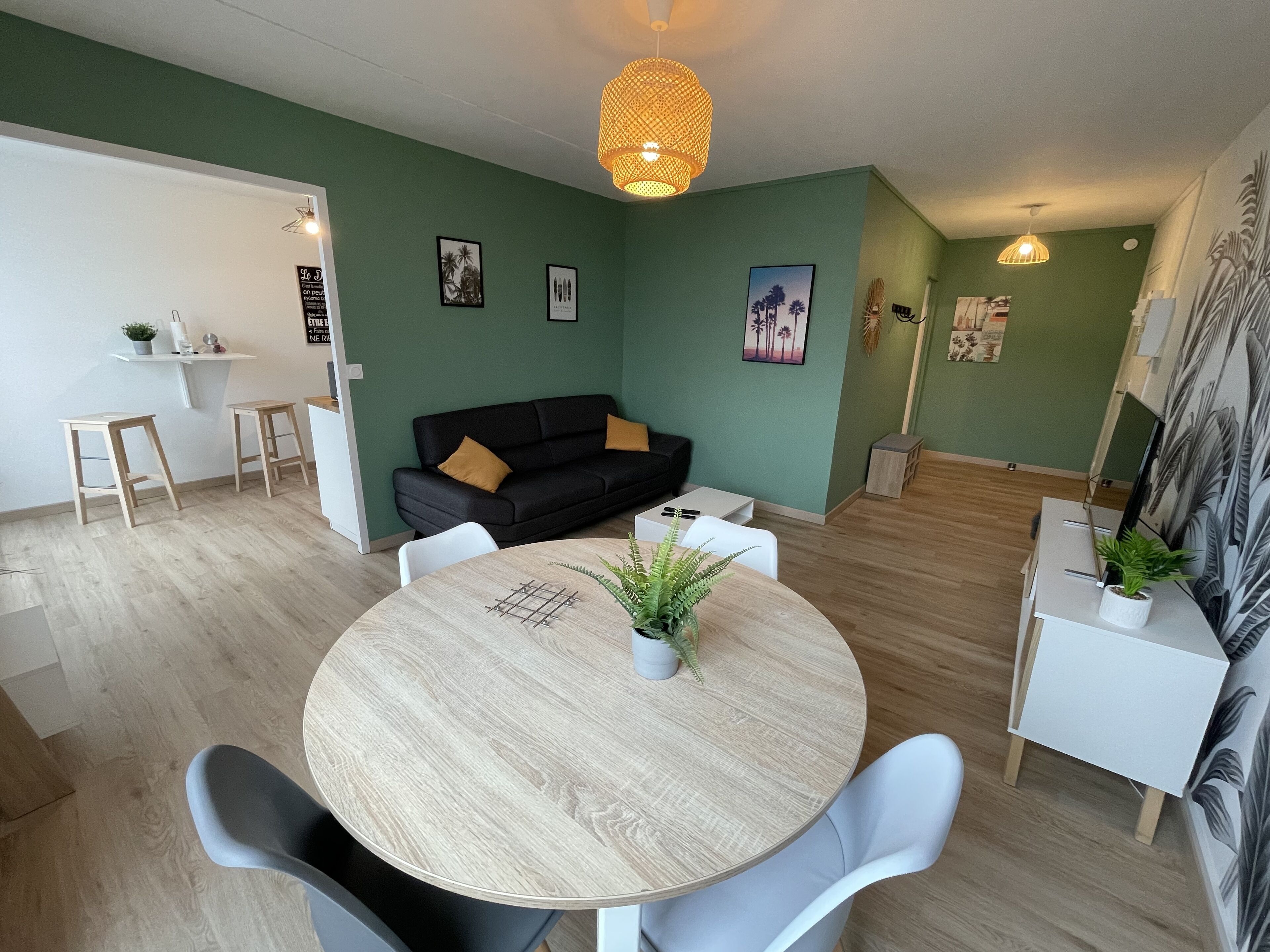 A modern living room with a round wooden table, a black sofa, and a white coffee table. There are two white chairs around the table, and a small plant on the table. The room has green walls and a wooden floor.