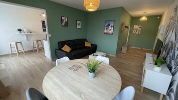 A modern living room with a round wooden table, a black sofa, and a white coffee table. There are two white chairs around the table, and a small plant on the table. The room has green walls and a wooden floor.