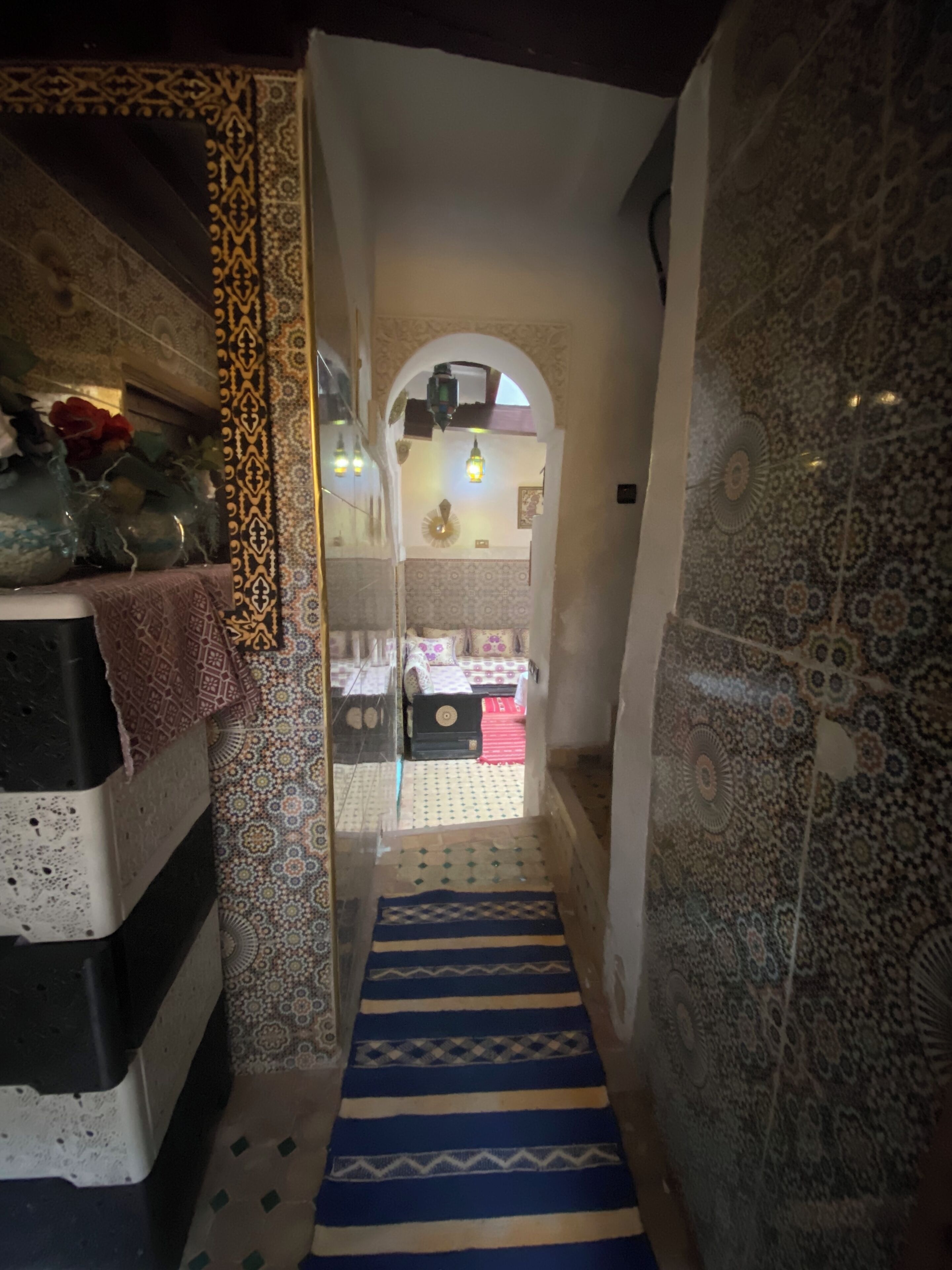 Un pasillo estrecho con paredes estampadas y una alfombra azul y blanca a rayas que conduce a una habitación con una cama y una pequeña mesa.