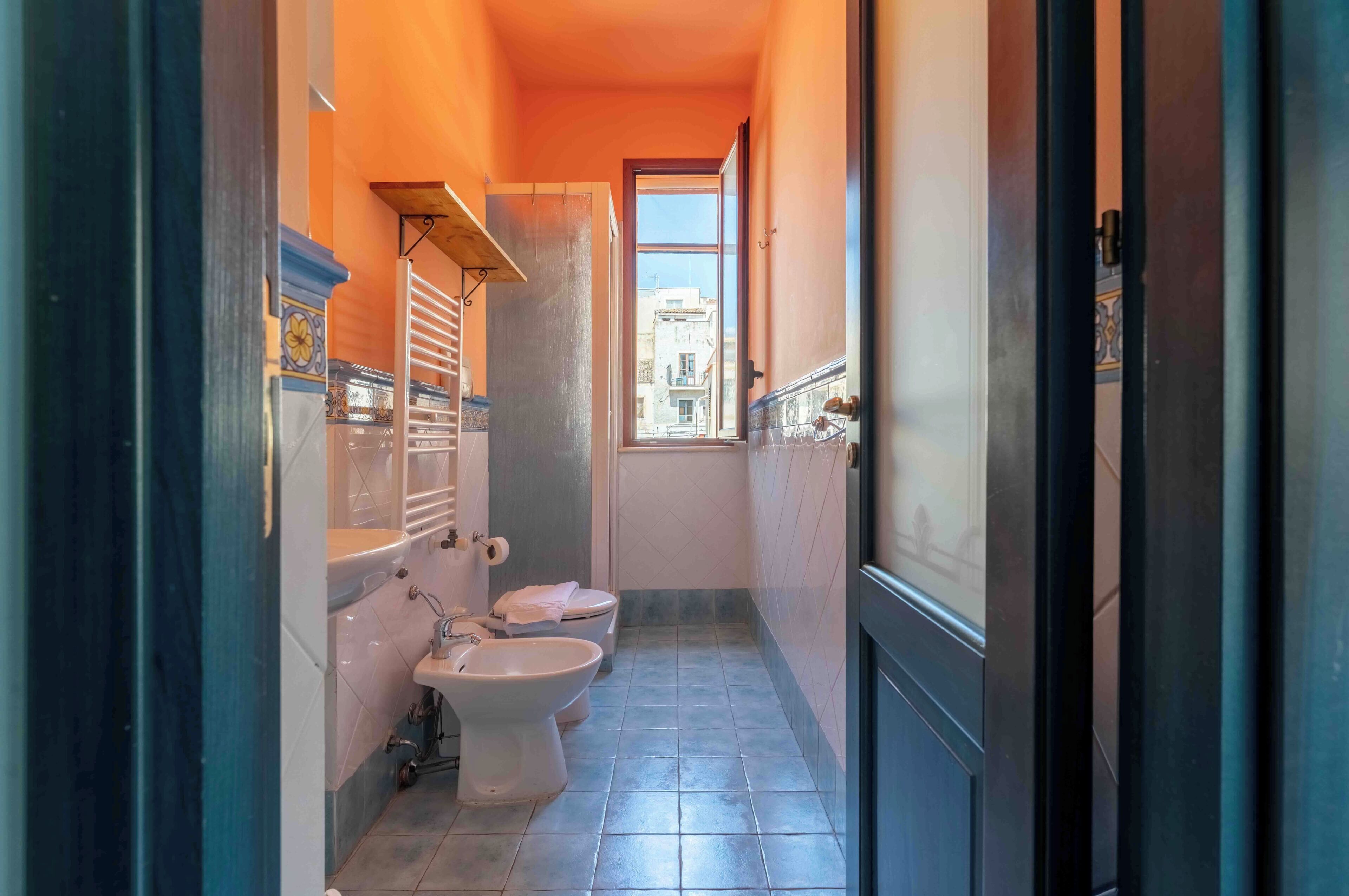 Un baño estrecho con inodoro, bidé y lavamanos, una ventana con vistas a edificios y suelo de baldosas.
