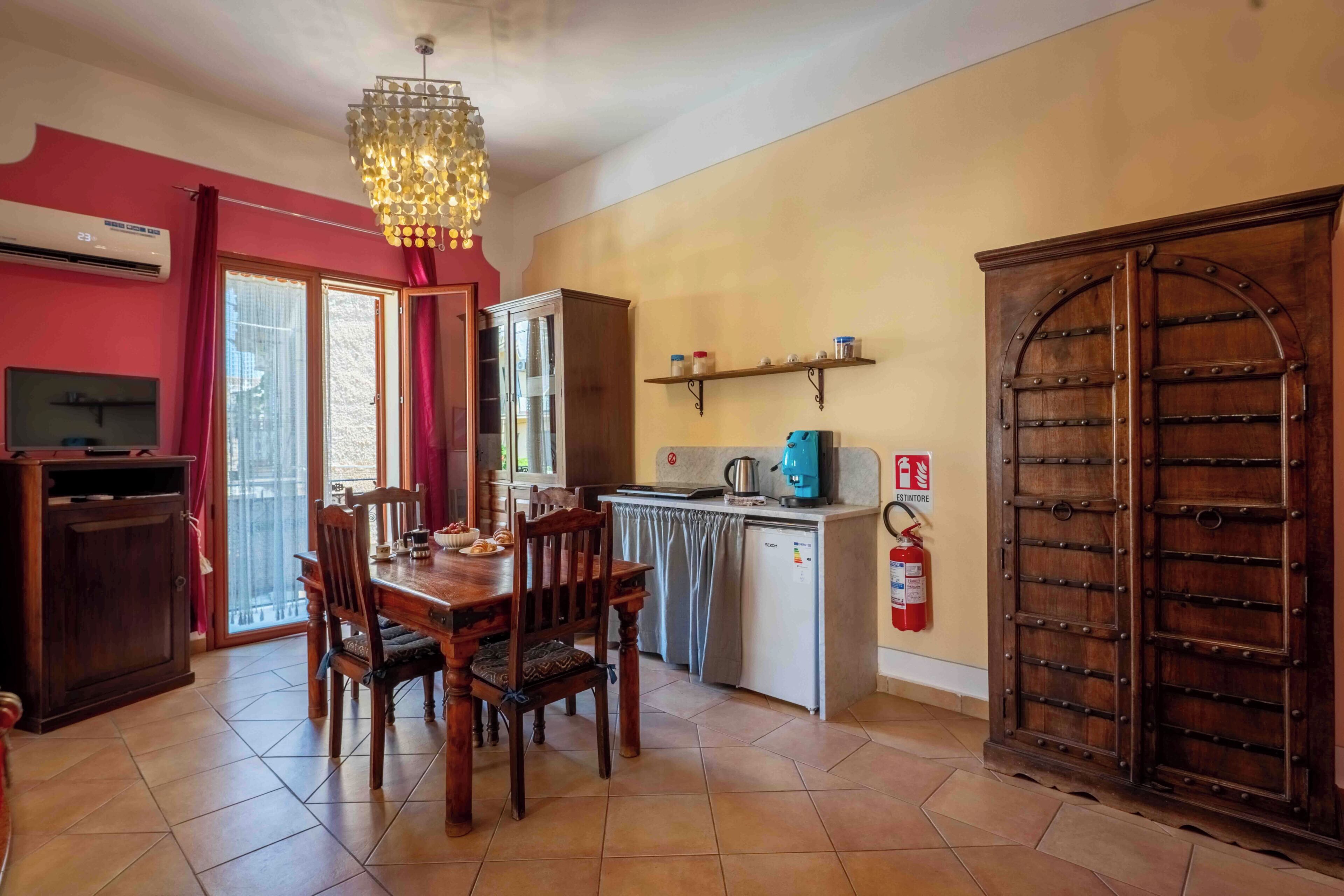 A dining area with a wooden table and chairs, a kitchen with a coffee maker, a fire extinguisher, and a large wooden door.