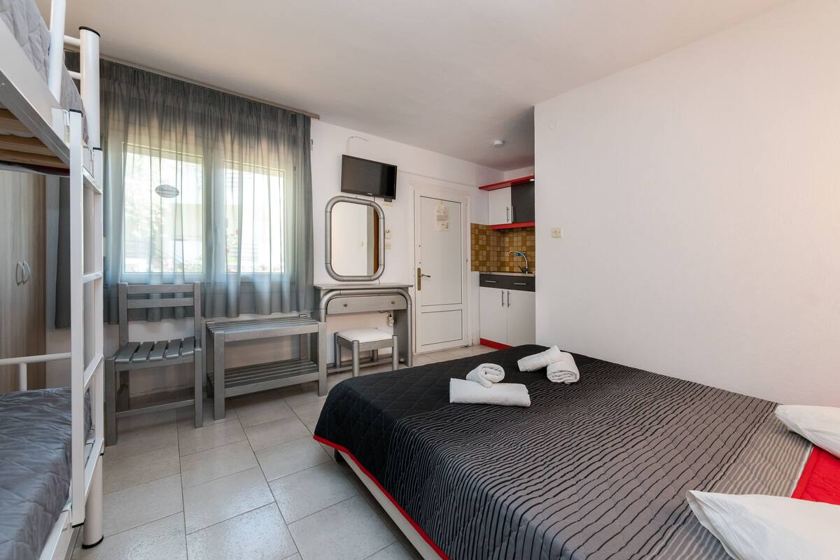 A hotel room with a bunk bed, a desk with a chair, a small table, a mirror, a television, a wardrobe, and a bed with towels.