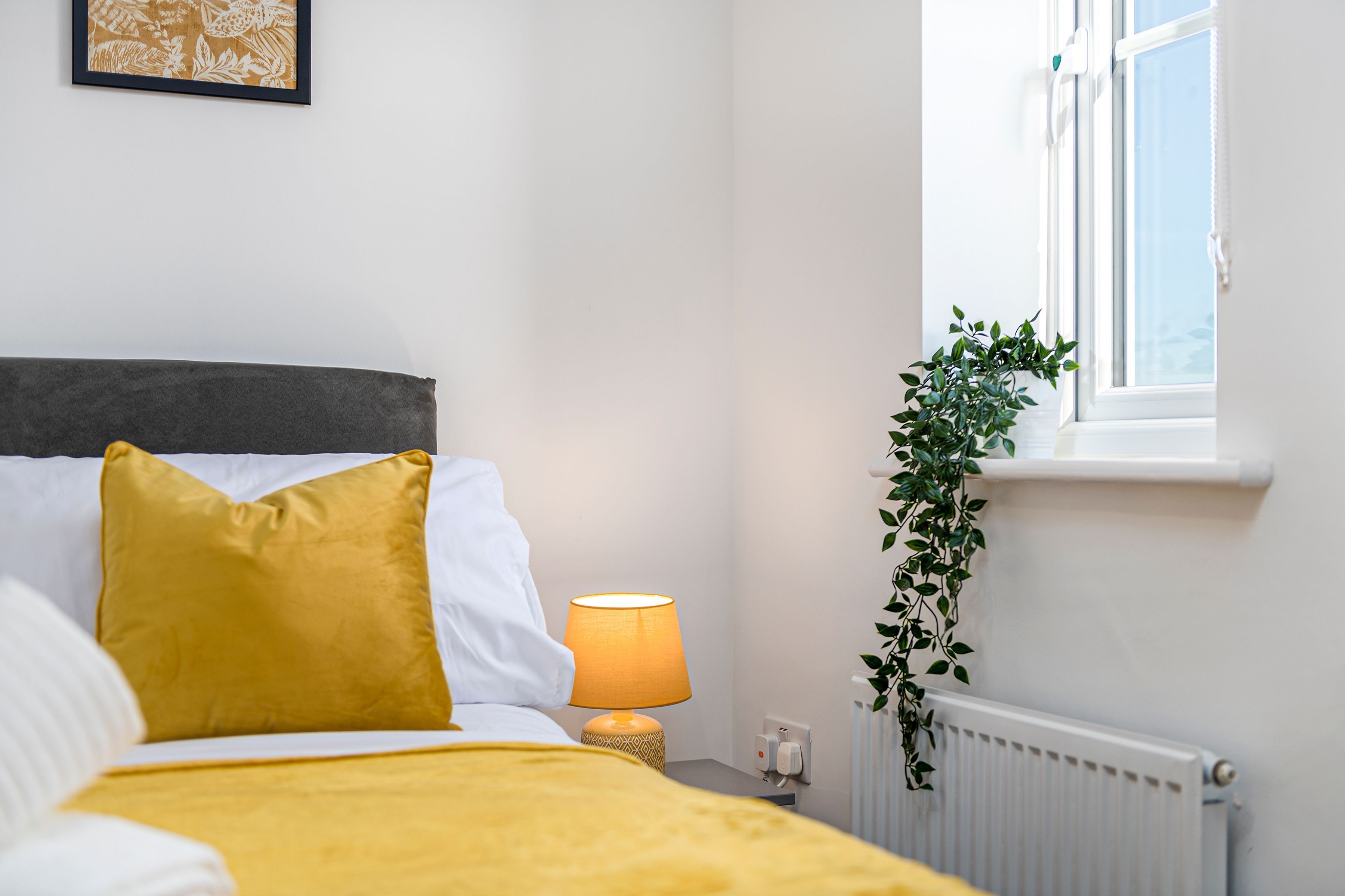 Una cama bien hecha con una manta y cojines amarillos, una lámpara de noche, un radiador y una ventana con una planta en el alféizar.