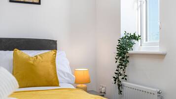 Una cama bien hecha con una manta y cojines amarillos, una lámpara de noche, un radiador y una ventana con una planta en el alféizar.