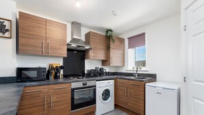 A modern kitchen with wooden cabinets, a built-in oven, a microwave, a washing machine, and a sink. - The Holt (Lincoln)